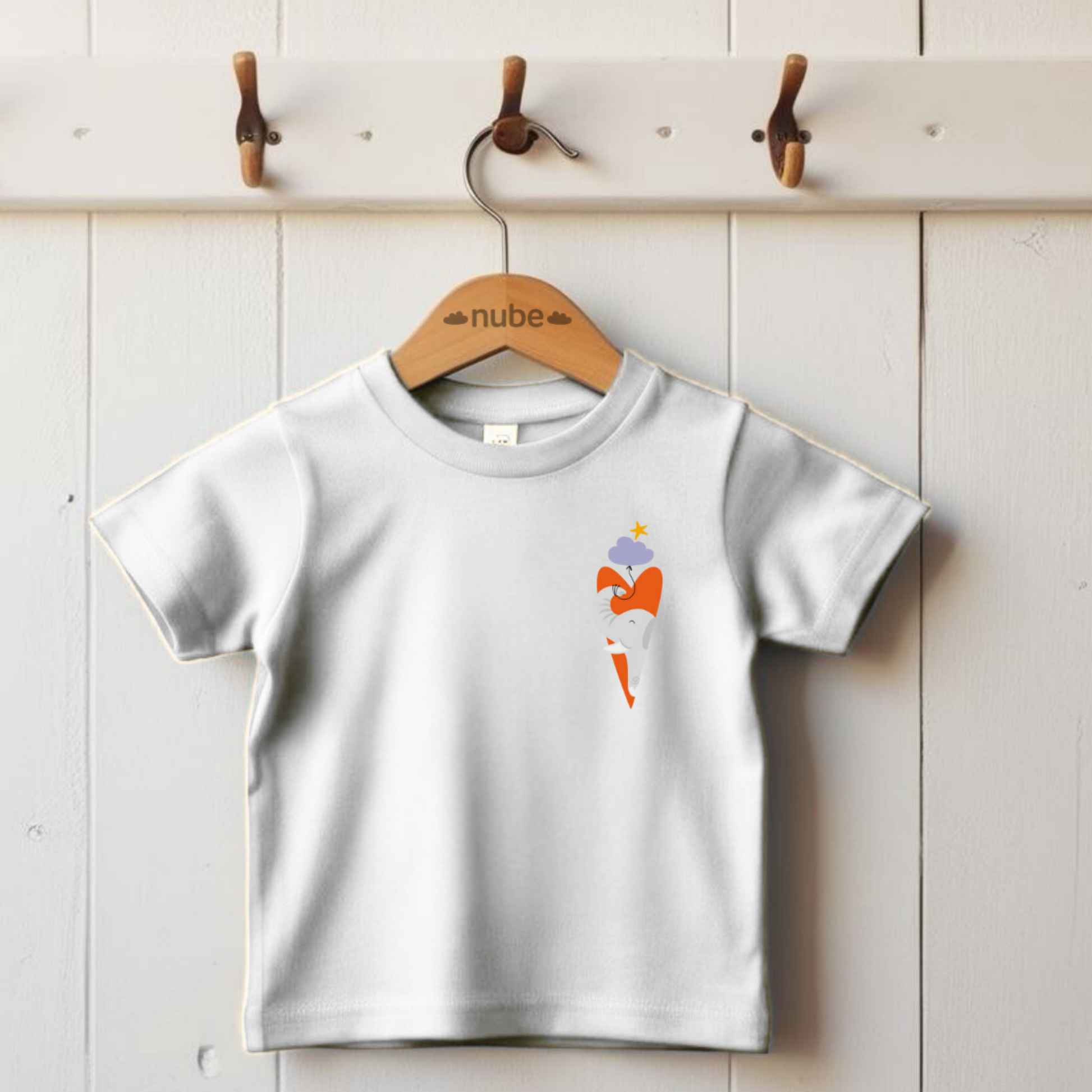 Playera blanca colgada en un gancho con diseño de un elefante gris dentro de un corazón naranja, con globo de nube morada amarrada a su trompa y una estrella amarilla en la parte superior del pecho. Arriba una estrella blanca lo guía