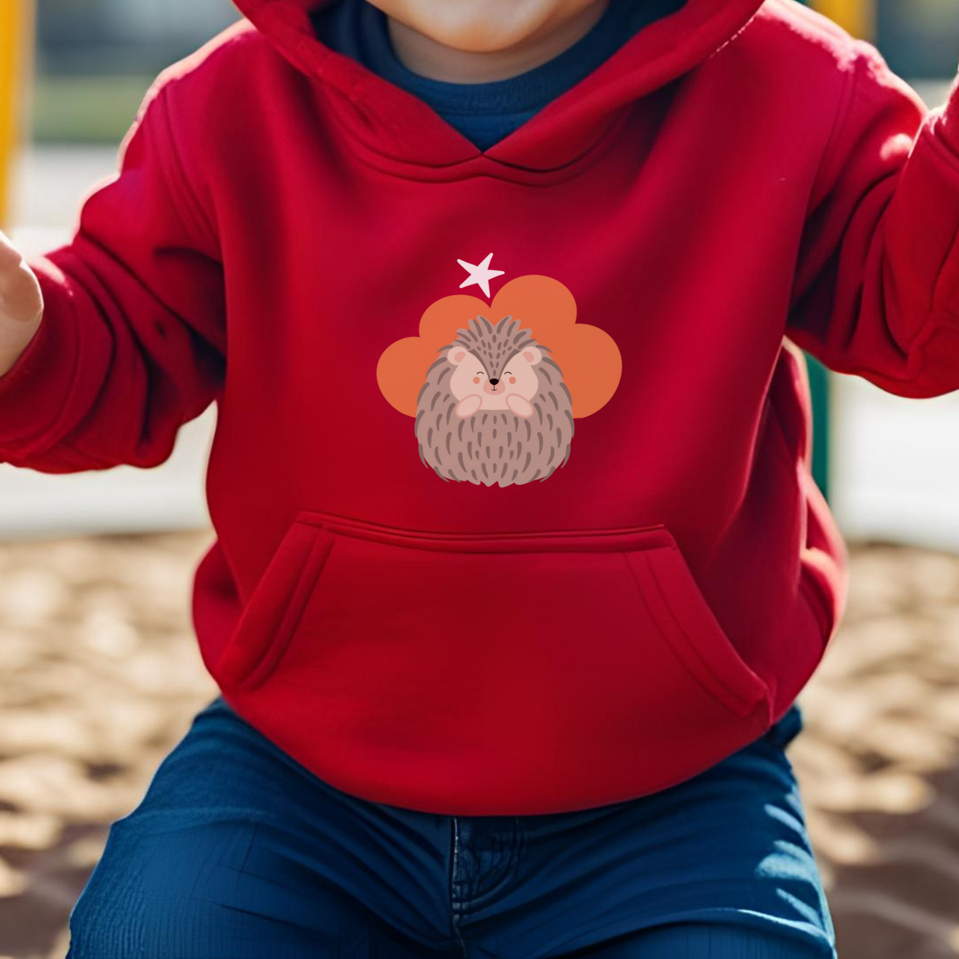 Toddler jugando en el parque con sudadera tipo hoodie roja con diseño de Filomena el Puercoespín de la marca de ropa Nube dentro de una nube naranja y una estrella blanca a lado.