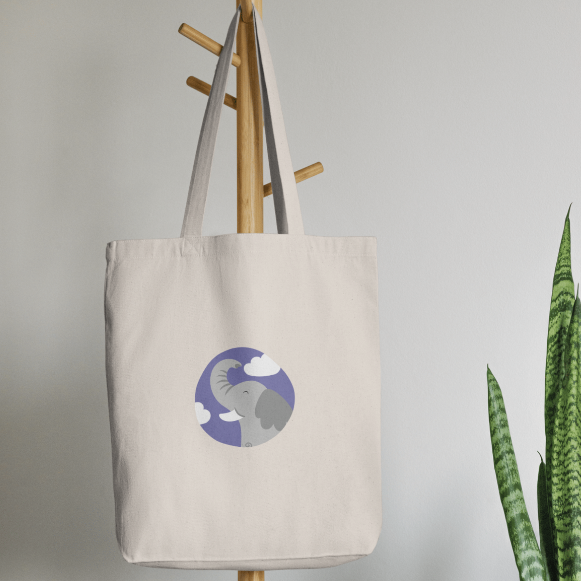 Bolsa Tote Bag color beige en  perchero, mostrando el retrato del elefante Clementino de la marca de ropa nube dentro de un círculo morado con nubes.