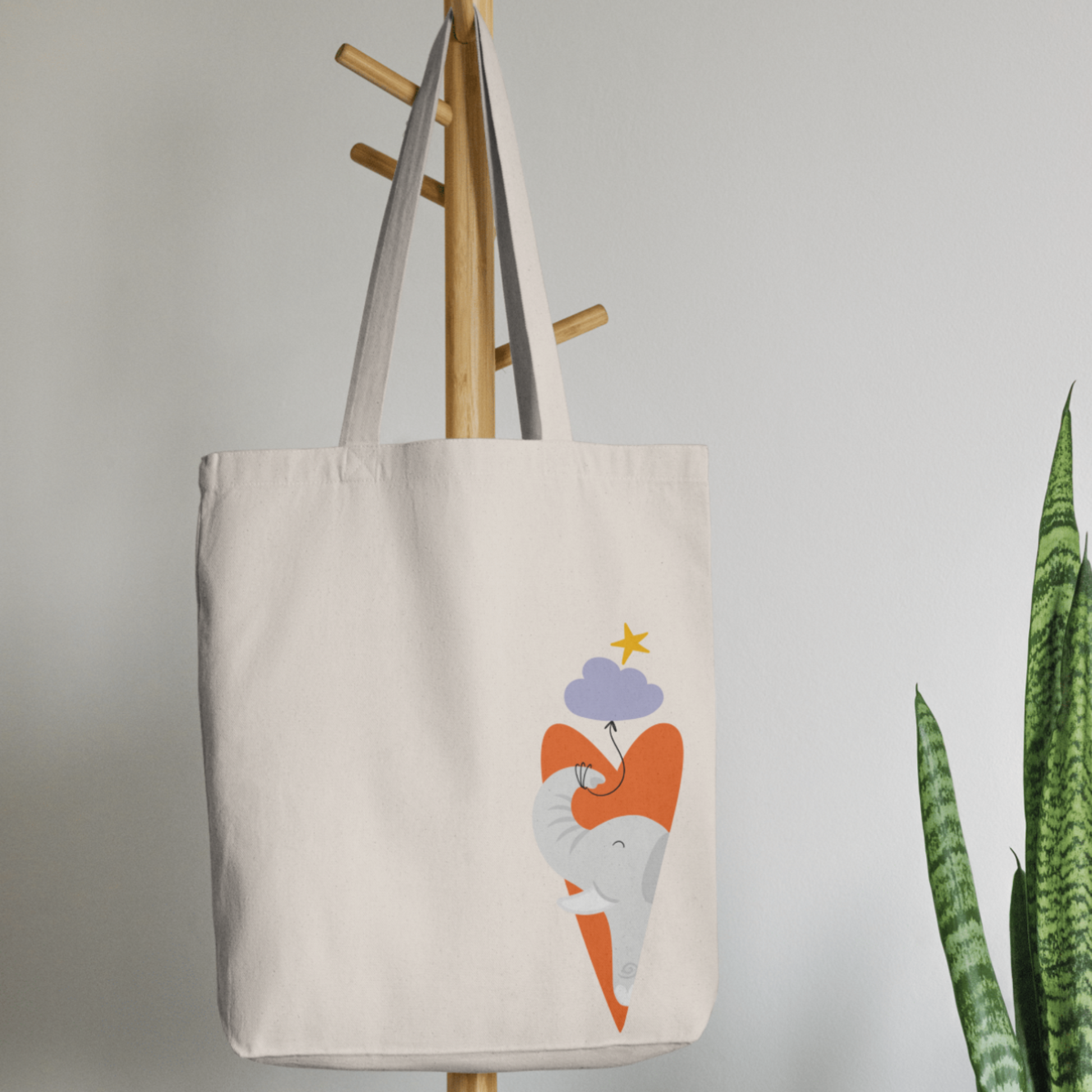 Bolsa Tote Bag color beige en  perchero, mostrando el diseño del elefante clementino de la marca de ropa nube dentro de un corazón naranja y con un globo morado amarrado en su trompa. Arriba los guía una estrella amarilla.