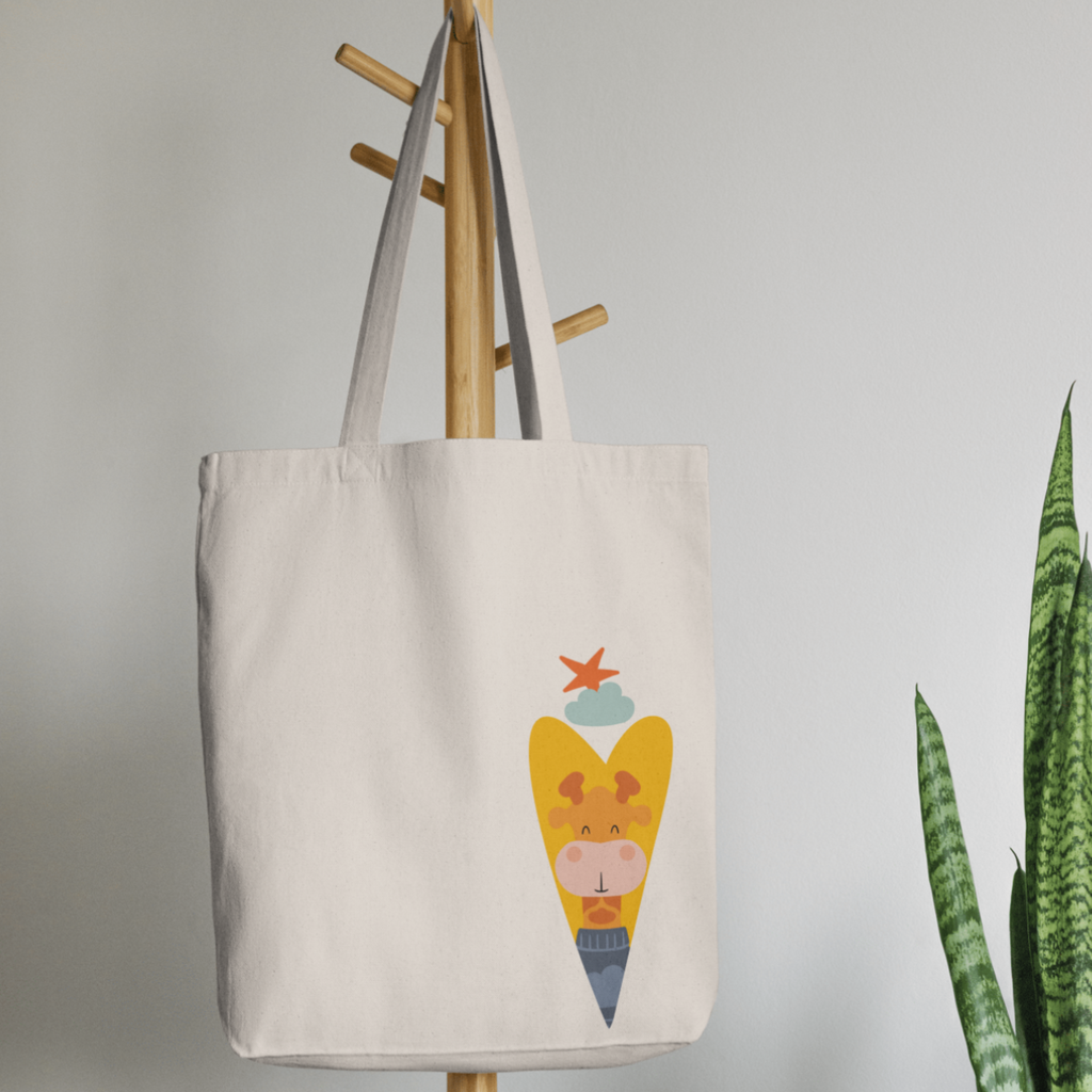 Bolsa Tote Bag color beige en  perchero, mostrando el diseño de la jirafa Lazaro de la marca de ropa nube dentro de un corazón amarillo. Arriba los guía una estrella naranja y una nube azul.