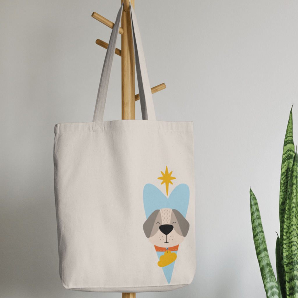Bolsa Tote Bag color beige en  perchero, mostrando el diseño del perro Leandro con collar naranja y placa amarilla de la marca de ropa nube dentro de un corazón azul. Arriba lo guía una estrella amarilla.
