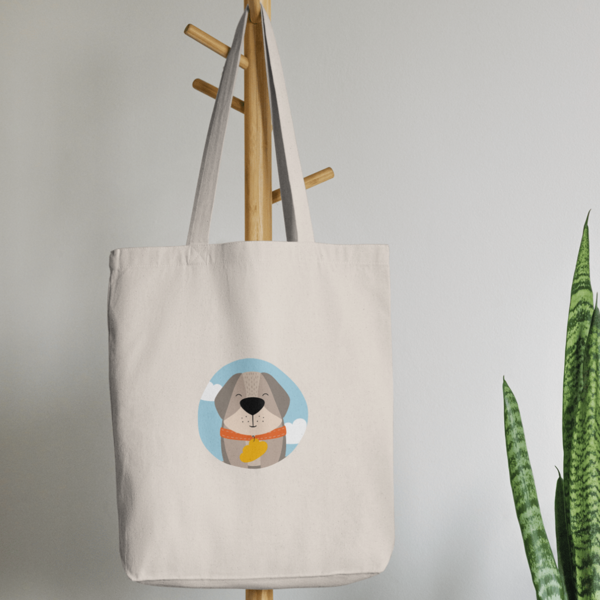 Bolsa Tote Bag color beige en  perchero, mostrando el retrato del perro Leandro con de la marca de ropa nube con collar naranja y placa amarilla en forma de nube dentro de un círculo azul con nubes.