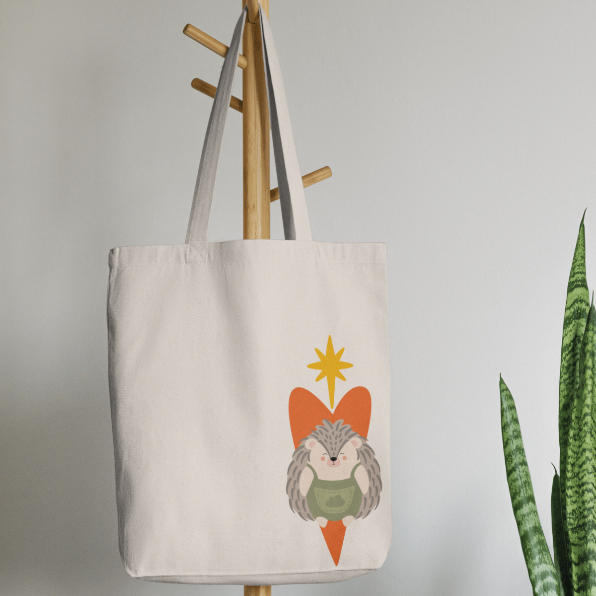Bolsa Tote Bag color beige en  perchero, mostrando el diseño del puercoespin Filomeno con un traje verde de la marca de ropa nube dentro de un corazón naranja Arriba lo guía una estrella naranja