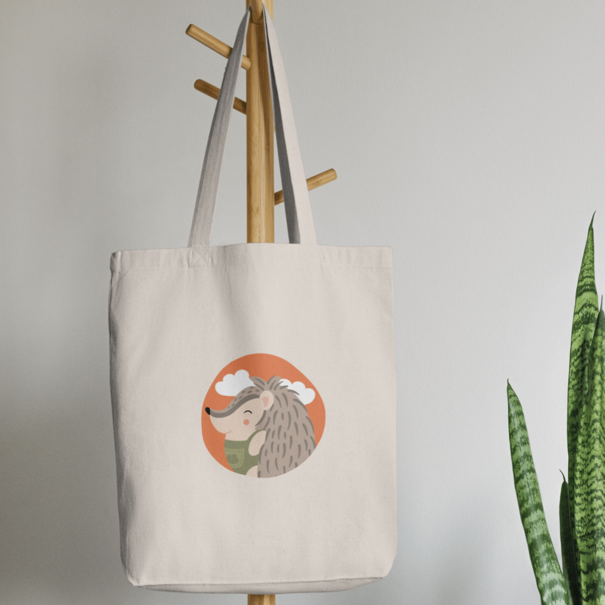 Bolsa Tote Bag color beige en  perchero, mostrando el retrato del puercoespin Filomeno con un traje verde de la marca de ropa nube dentro de un círculo naranja con nubes.